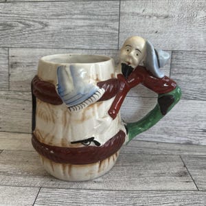 May include: A vintage ceramic beer mug with a whimsical design. The mug features a handle shaped like a man in a red coat and green pants. The mug's body is cream-colored with brown and white accents.