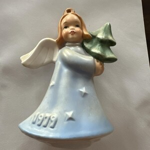 May include: A ceramic angel Christmas ornament. The angel wears a light blue dress with star accents and holds a green Christmas tree. The ornament has a loop for hanging and the year "1979" is printed on the dress.