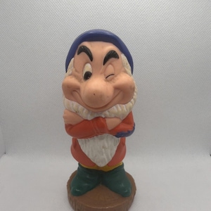 May include: A moulded plastic figurine of the character Grumpy from Snow White, featuring a blue cap, red jacket, and green boots. The character has a white beard, crossed arms, and is winking with one eye. It stands on a brown circular base.