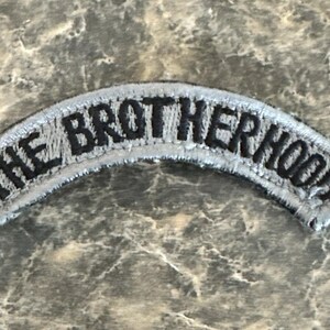 May include: A light gray, arched patch embroidered with the words "THE BROTHERHOOD" in black block letters. The patch has a light gray border and is set against a mottled gray background.