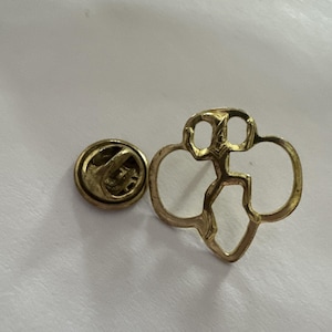 May include: A gold-colored Girl Scout pin with a pin back. The pin features the Girl Scout trefoil logo, a stylized figure within three connected circles. The pin is made of metal and has a shiny finish.