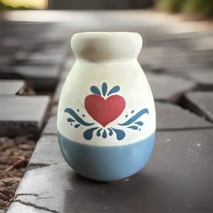 May include: A small, hand-painted ceramic vase with a cream-colored top and a blue base. The vase features a red heart design with blue floral accents. The vase is a decorative item.