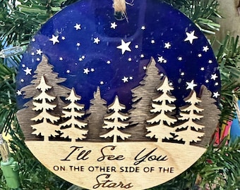 May include: Round Christmas ornament with a dark blue background and white stars. The ornament features a wooden forest silhouette and the text "I'll See You on the Other Side of the Stars". A natural twine loop is attached for hanging.