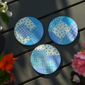 May include: Three circular coasters featuring a blue and white patchwork design. Each coaster displays a variety of patterns including small floral prints, checkered squares, and dotted textures in shades of light and dark blue on a white background.