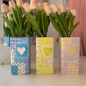 May include: Three rectangular vases with patchwork designs in blue, yellow, and pink, each featuring a heart applique. The vases hold tulips with pale yellow petals and green stems, creating a charming floral arrangement.