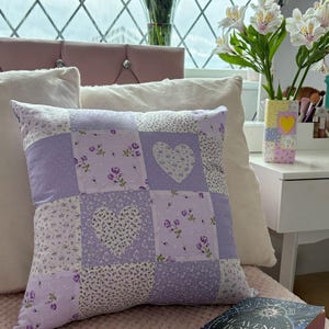 Puede incluir: Un cojín decorativo cuadrado de patchwork con una cuadrícula de cuadrados de tela en color lavanda y blanco. El diseño incluye patrones alternos de pequeños estampados florales morados, lunares blancos y dos aplicaciones de tela en forma de corazón.