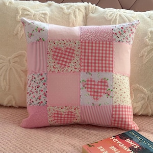 May include: A pink patchwork cushion with floral, gingham, and striped patterns. Two squares feature heart shapes. The cushion rests on a pink bedspread, with a book titled "Where the Crawdads Sing" visible.