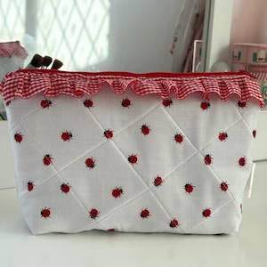 May include: White quilted cosmetic bag with a red gingham ruffle trim and ladybug pattern. The bag has a zipper closure and a diamond pattern. The bag is sitting on a white surface.