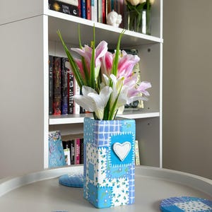 May include: A blue and white patchwork vase with a white heart applique, holding pink and white lilies. The vase sits on a round table with matching coasters. A white bookshelf with books and flowers is in the background.