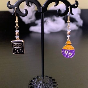 May include: A pair of gold-toned earrings with a Halloween theme. One earring features a black book with the word "MAGIC" and a crescent moon. The other earring has a purple potion bottle with two eyes and an orange top.