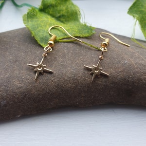 May include: A pair of gold star earrings with a simple design. The earrings are hanging from a gold hook.
