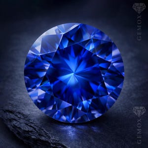 May include: A round, faceted, brilliant-cut sapphire gemstone. The gem is a deep, vibrant blue color, with multiple facets reflecting light. The gemstone is set against a dark, textured background. The GEMOX logo is visible on the right side.