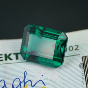 K&ouml;nnte beinhalten: Ein rechteckiger, smaragdgeschliffener Edelstein mit tiefgr&uuml;ner Farbe. Der Edelstein hat eine facettierte Oberfl&auml;che, die das Licht reflektiert und einen brillanten Glanz erzeugt. Der Edelstein liegt vor einem hellen Hintergrund mit Text und einem Logo.