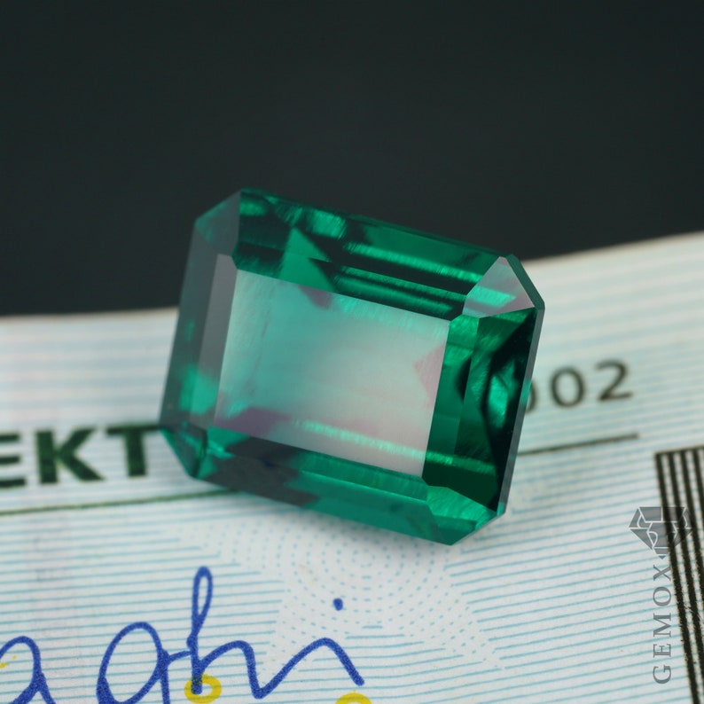K&ouml;nnte beinhalten: Ein rechteckiger, smaragdgeschliffener Edelstein mit tiefgr&uuml;ner Farbe. Der Edelstein hat eine facettierte Oberfl&auml;che, die das Licht reflektiert und einen brillanten Glanz erzeugt. Der Edelstein liegt vor einem hellen Hintergrund mit Text und einem Logo.