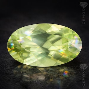 Pode incluir: Uma pedra preciosa oval, verde claro, com facetas internas que refletem um arco-íris de cores. A gema está sobre um fundo escuro, realçando sua clareza e brilho. É um peridoto.
