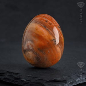 May include: A polished, egg-shaped stone with swirling patterns of orange, brown, and cream. The gemstone has a smooth, glossy surface and is displayed against a dark background. The egg shape is a classic form, often used for decorative purposes.