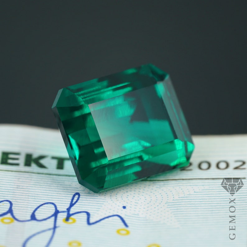 K&ouml;nnte beinhalten: Ein facettierter, smaragdgeschliffener Edelstein in tiefgr&uuml;ner Farbe. Der Stein ist hochglanzpoliert und reflektiert das Licht, wodurch seine innere Struktur sichtbar wird. Der Hintergrund ist unscharf.