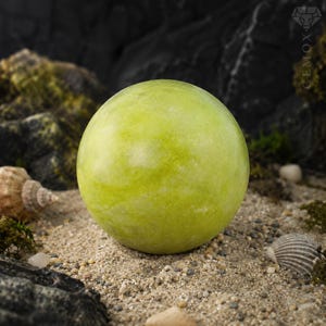 Può includere: Una gemma sferica verde chiaro. La pietra è posta su una superficie sabbiosa con piccole rocce, conchiglie e muschio sullo sfondo. La gemma è il punto focale dell'immagine.