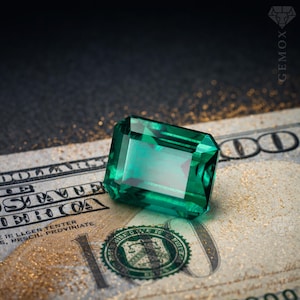 K&ouml;nnte beinhalten: Ein rechteckiger, smaragdgr&uuml;ner Edelstein liegt auf einem Hundert-Dollar-Schein. Die Facetten des Edelsteins reflektieren das Licht und zeigen seine Klarheit. Der Hintergrund ist ein dunkler Farbverlauf mit dezentem Goldglitter.