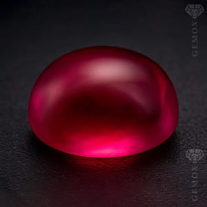 Może przedstawiać: Wypolerowany kamień szlachetny rubinu w kształcie kaboszonu. Powierzchnia odbija światło, ukazując głęboki czerwony odcień z subtelnym blaskiem. Kamień szlachetny umieszczony na ciemnym, teksturowanym tle.