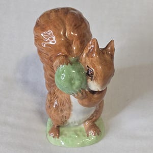 May include: A brown and white ceramic squirrel figurine holding a green acorn. The squirrel is standing on a green base.