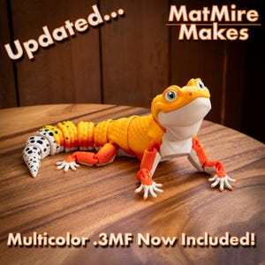 May include: An articulated gecko figurine featuring a vibrant orange and yellow body with a white underbelly and a white tail with black spots. The text "Updated... MatMire Makes" and "Multicolor .3MF Now Included!" is visible in the image.