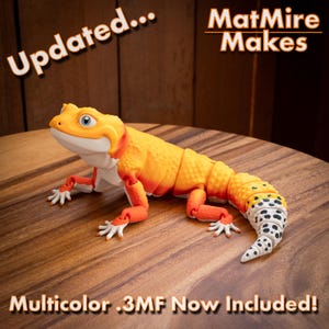 May include: A 3D printed articulated lizard figurine featuring a vibrant orange and yellow gradient body with a white underbelly and a white tail patterned with black spots. The text "Updated... MatMire Makes" and "Multicolor .3MF Now Included!" appears.
