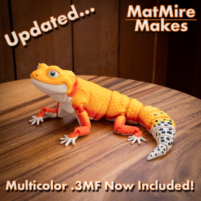 May include: A 3D printed articulated lizard figurine featuring a vibrant orange and yellow gradient body with a white underbelly and a white tail patterned with black spots. The text "Updated... MatMire Makes" and "Multicolor .3MF Now Included!" appears.