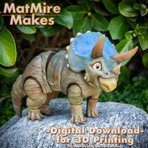 Op de afbeelding: Een 3D-geprint model van een groene en blauwe triceratops-dinosaurus met een tandenrij. Het model staat op een rots met groen gebladerte op de achtergrond. De tekst "MatMire Makes - Digital Download for 3D Printing" staat onderaan de afbeelding.