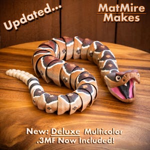 May include: A segmented, articulated toy rattlesnake with a brown, tan, and white patterned body, an open mouth with visible white fangs, and a pink interior. Text reads: Updated... MatMire Makes. New: Deluxe Multicolor .3MF Now Included!