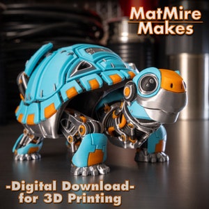 Puede incluir: Un modelo de tortuga robótica impreso en 3D con un caparazón azul y naranja. El modelo está diseñado para la impresión 3D y está disponible para descarga digital. El texto "MatMire Makes" es visible en el fondo.