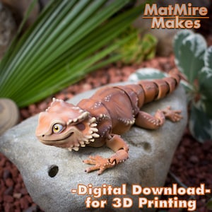 May include: A 3D printed model of a brown and orange lizard with articulated joints. The lizard is posed on a rock with a green plant in the background. The text "MatMire Makes - Digital Download - for 3D Printing" is printed on the image.