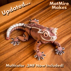 Puede incluir: Una figura de gecko articulada en tonos marrón, beige y blanco con ojos naranjas. El texto "Updated... MatMire Makes" y "Multicolor .3MF Now Included!" es visible. El gecko tiene una cola y extremidades segmentadas.