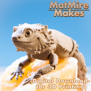 May include: A 3D printed model of a lizard, with a light brown color, sitting on a rock. The lizard has a friendly expression and is posed in a sitting position. The text "MatMire Makes - Digital Download - for 3D Printing" is visible at the top of the image.