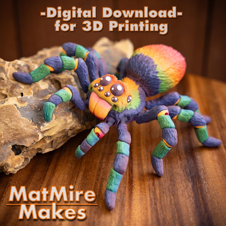 May include: A colorful 3D printed spider model with a rainbow color scheme. The spider has a large, round head with googly eyes and a long, orange tongue. The spider is sitting on a rock.