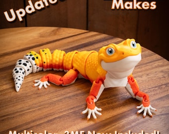 May include: An articulated gecko figurine featuring a gradient orange and yellow body with a white underbelly and a tail patterned with black spots. The text "Updated... MatMire Makes" and "Multicolor .3MF Now Included!" is visible in the image.