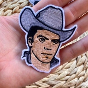 May include: Embroidered patch featuring a cowboy's face with a gray hat and a tan complexion. The patch has a white border and is held in a person's hand. The background is a woven, light brown surface.