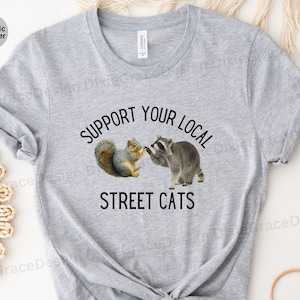 May include: A heather grey t-shirt with the text "Support Your Local Street Cats" printed in black. The shirt features a graphic of a raccoon and a squirrel shaking hands.