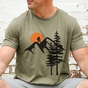 May include: A green t-shirt with a graphic of a Bigfoot walking on a mountain range with a sunset behind him. Two pine trees are in the foreground.