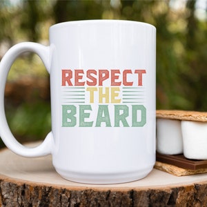 May include: White ceramic coffee mug with the text "Respect the Beard" in a retro style font with red, yellow, and green colors. The mug is sitting on a wooden surface.