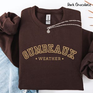 May include: A dark chocolate-colored sweatshirt with the text "GUMBEAUX WEATHER" in a gold, collegiate-style font. A silver necklace with a sun pendant is draped across the front. The sweatshirt is folded on a white surface.