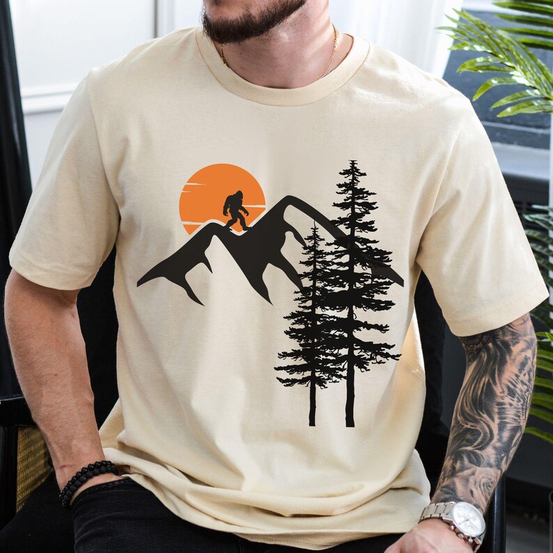 May include: A beige t-shirt with a black silhouette of a Bigfoot walking on a mountain range with a sunset behind him. Two black trees are in the foreground.