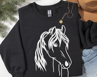 May include: Black sweatshirt featuring a white horse head graphic. The word "Black" is printed at the top. A gold necklace with a pendant is draped over the sweatshirt. A brown hat and denim jeans are in the background.