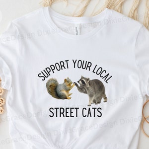 May include: White t-shirt with a black graphic that says "Support Your Local Street Cats". The graphic features a raccoon and a squirrel.