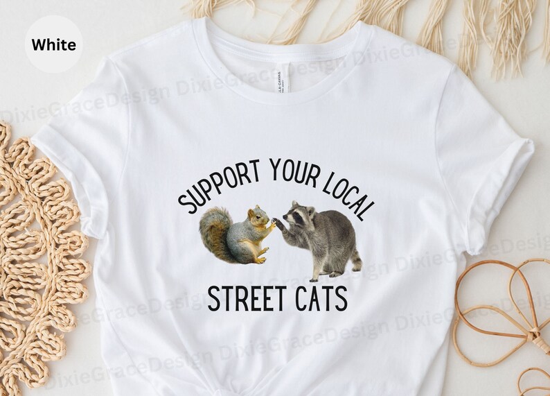 May include: White t-shirt with a black graphic that says "Support Your Local Street Cats". The graphic features a raccoon and a squirrel.