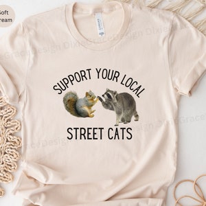 May include: A cream coloured t-shirt with the text "Support Your Local Street Cats" in black. A cartoon squirrel and raccoon are shaking hands above the text.