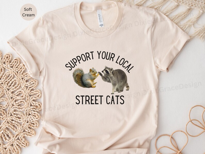 May include: A cream coloured t-shirt with the text "Support Your Local Street Cats" in black. A cartoon squirrel and raccoon are shaking hands above the text.