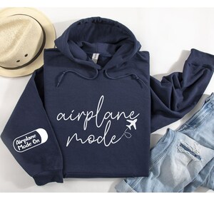 Peut inclure: Un sweat &agrave; capuche bleu marine avec un motif blanc qui dit "mode avion" avec une petite ic&ocirc;ne d'avion. La manche gauche a un patch blanc qui dit "Mode Avion Activ&eacute;".