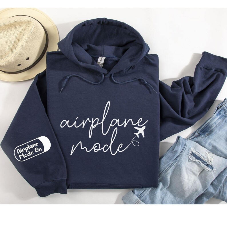 Peut inclure: Un sweat &agrave; capuche bleu marine avec un motif blanc qui dit "mode avion" avec une petite ic&ocirc;ne d'avion. La manche gauche a un patch blanc qui dit "Mode Avion Activ&eacute;".