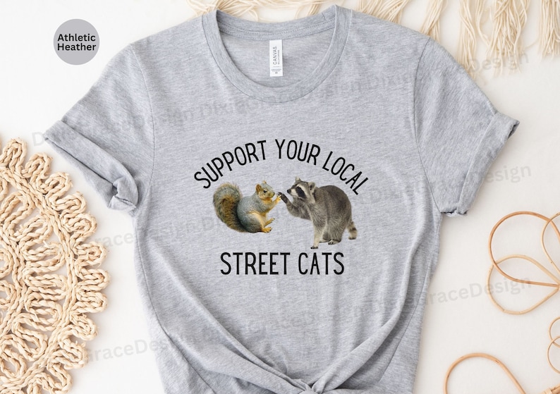 May include: A heather grey t-shirt with the text "Support Your Local Street Cats" printed in black. The shirt features a graphic of a raccoon and a squirrel shaking hands.